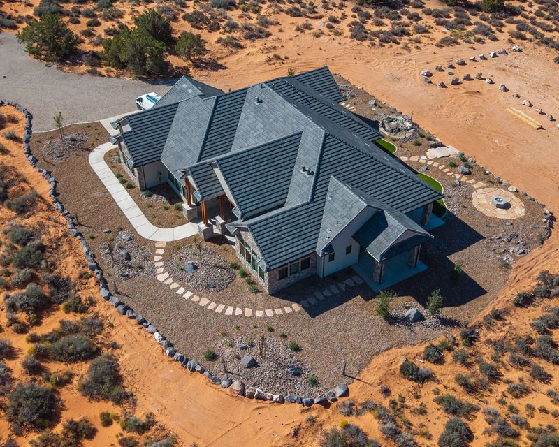 Aerial view of a custom desert home with a boulder border, desert plantings, stepping-stone paths, a flagstone fire pit patio, and some turf